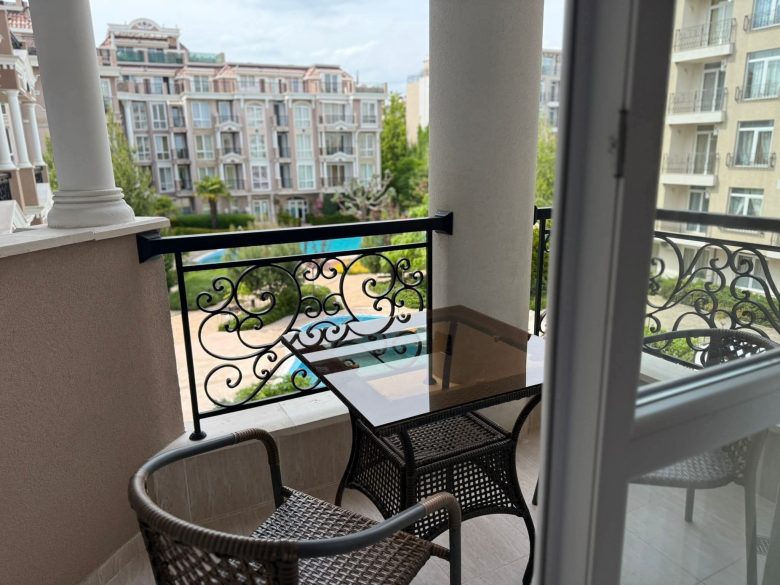 A small balcony with two wicker chairs and a glass-top table overlooks an apartment complex courtyard with greenery and a swimming pool, seen through an open glass door.