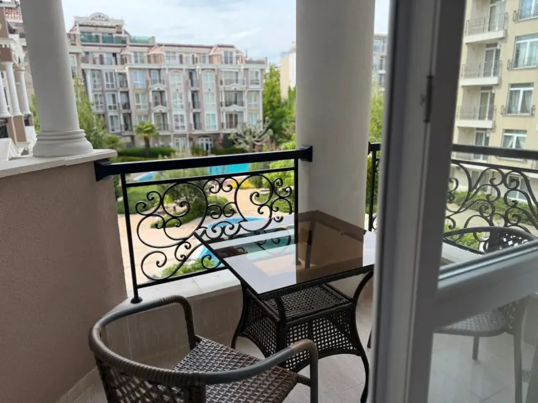 A small balcony with two wicker chairs and a glass-top table overlooks an apartment complex courtyard with greenery and a swimming pool, seen through an open glass door.