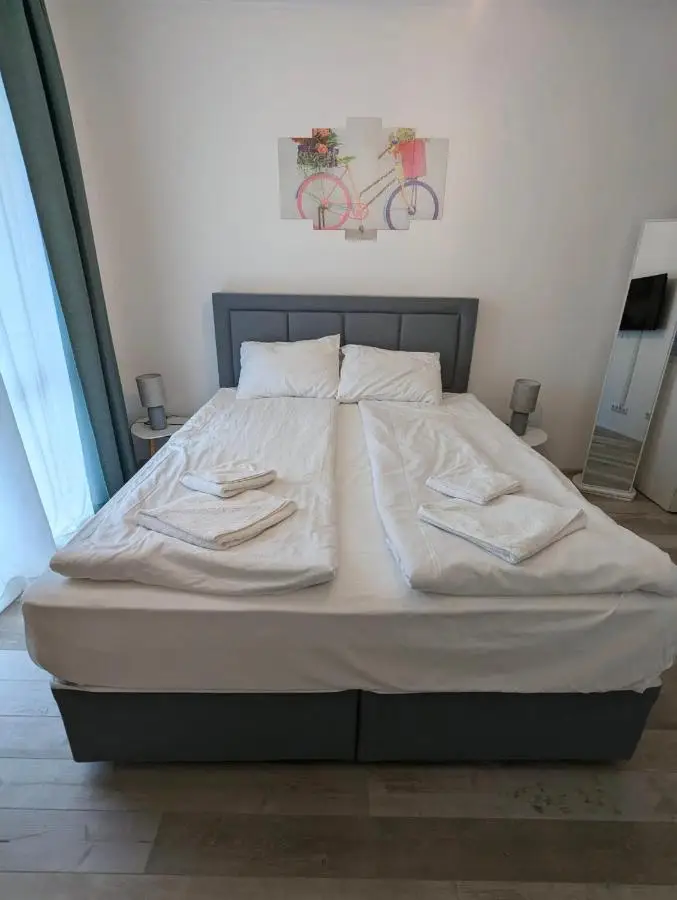 A neatly made bed with two pillows and folded white towels arranged on top, set in a modern, minimalistic bedroom with light-colored walls and a bicycle-themed artwork above the headboard.