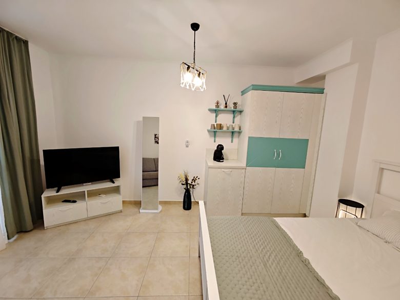 A modern, tidy bedroom with a bed, white wardrobe with teal accents, TV on a white stand, standing mirror, shelves with decor, and light-colored tile floor. Curtains hang beside a large window out of view.