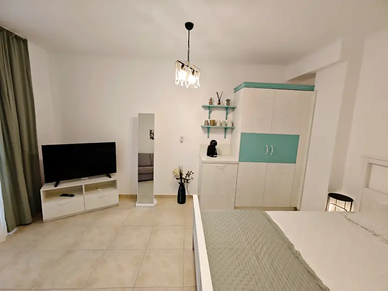 A modern, tidy bedroom with a bed, white wardrobe with teal accents, TV on a white stand, standing mirror, shelves with decor, and light-colored tile floor. Curtains hang beside a large window out of view.