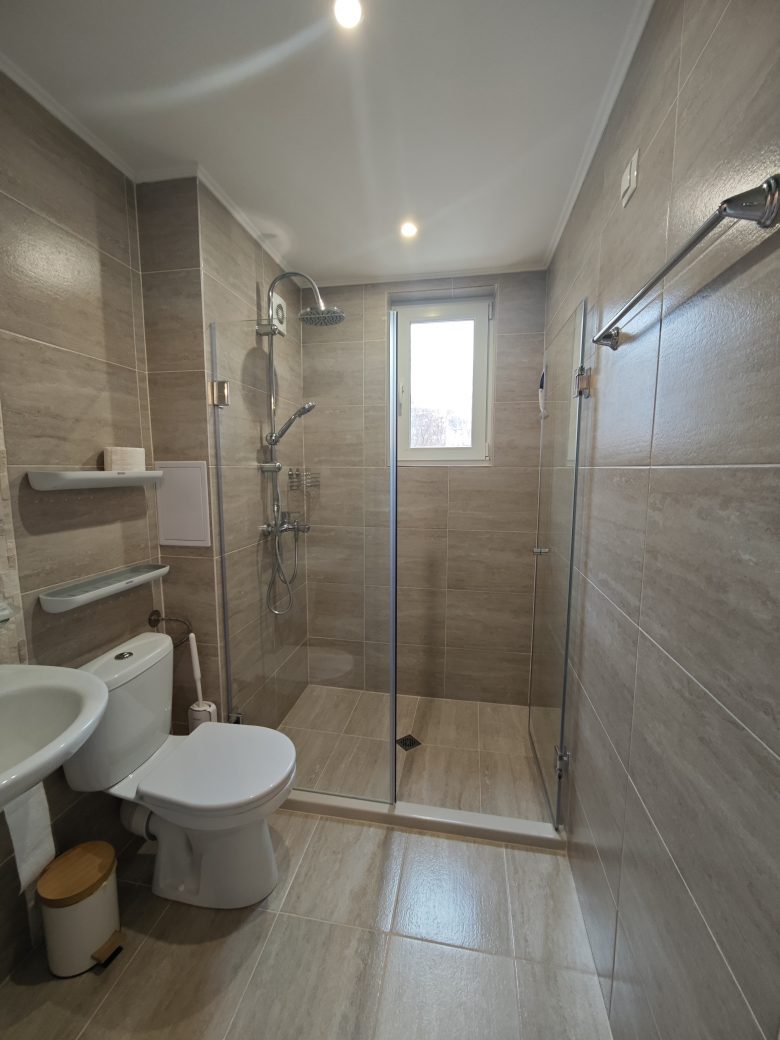 Modern bathroom with beige tiles, a walk-in glass shower, a white toilet, a white sink, shelves with toiletries, a towel rack, and a small window letting in natural light.