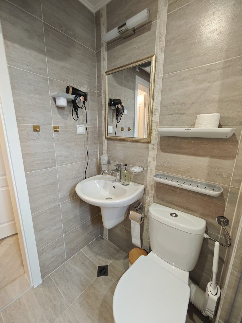 Modern bathroom with beige tiles, a wall-mounted sink, mirror, hairdryer, shelves with toiletries, a toilet, and a roll of toilet paper. The space is compact and neatly organized.