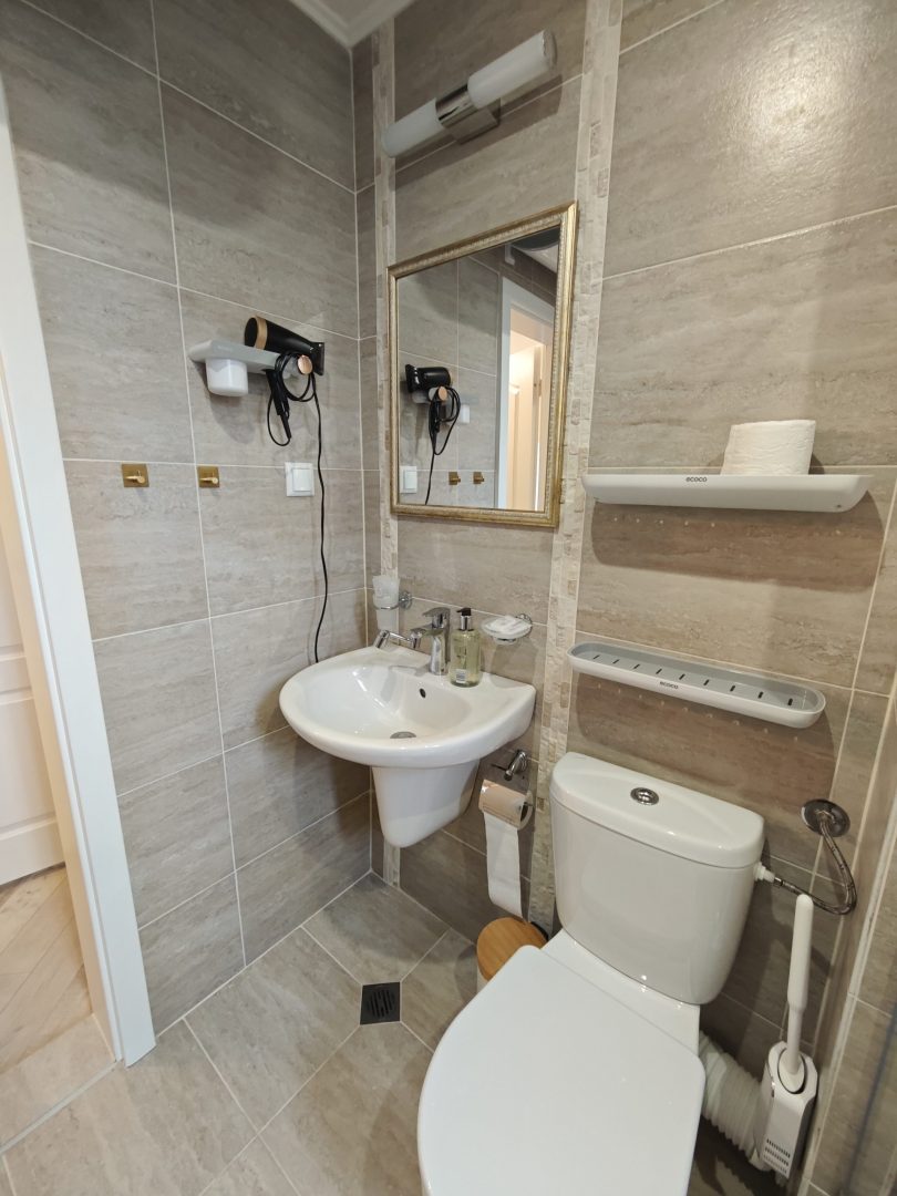 Modern bathroom with beige tiles, a wall-mounted sink, mirror, hairdryer, shelves with toiletries, a toilet, and a roll of toilet paper. The space is compact and neatly organized.