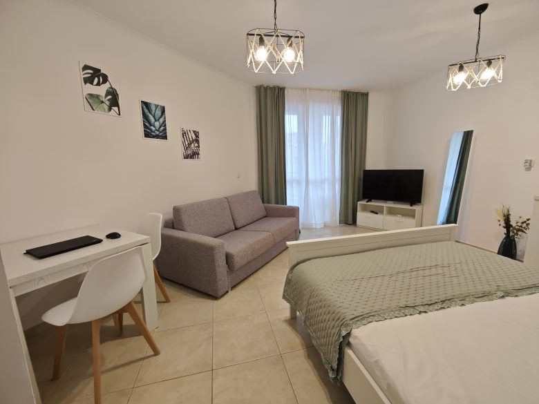 A modern, minimalistic bedroom with a bed, gray sofa, wall-mounted TV, small desk and chair, large mirror, and green curtains over a window. Light fixtures and decorative wall art add to the cozy ambiance.