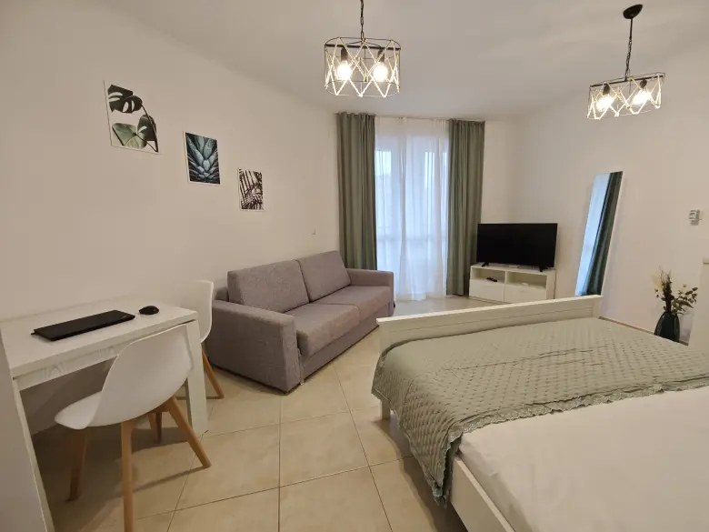 A modern, minimalistic bedroom with a bed, gray sofa, wall-mounted TV, small desk and chair, large mirror, and green curtains over a window. Light fixtures and decorative wall art add to the cozy ambiance.