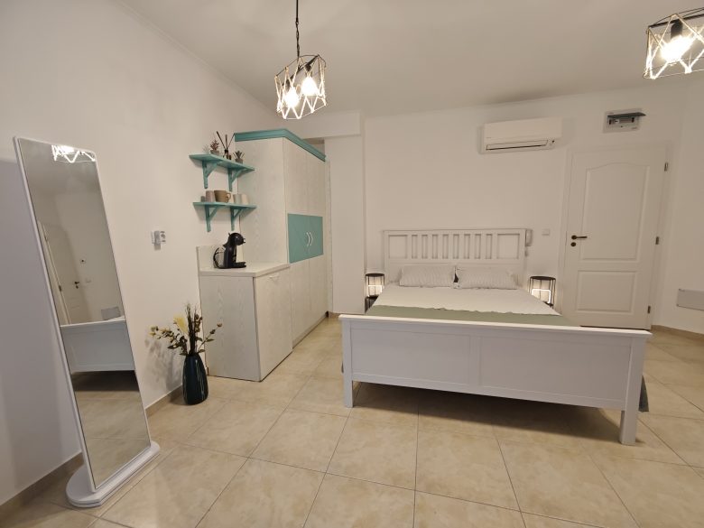 A modern, minimalistic bedroom with a white bed, bedside lamps, a standing mirror, a cabinet with shelves and a coffee machine, tiled floor, and white walls with subtle teal accents.