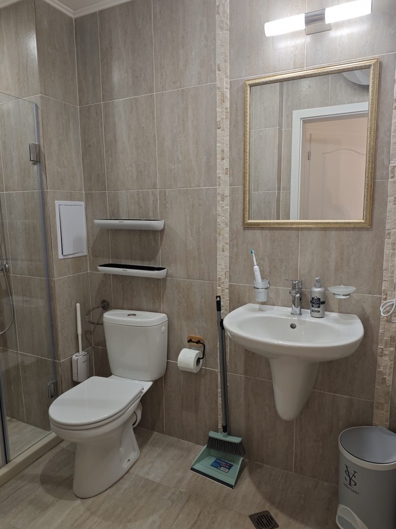 Modern bathroom with beige tiled walls, a white toilet, a wall-mounted sink with toiletries, a mirror above the sink, shelves, a toilet brush, a mop, a trash bin, and a shower area with a glass door.