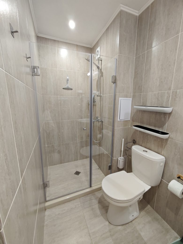 Modern bathroom with beige tiles, a glass-enclosed shower on the left, a white toilet on the right, and wall-mounted shelves above the toilet. The space is clean and well-lit with a ceiling light.