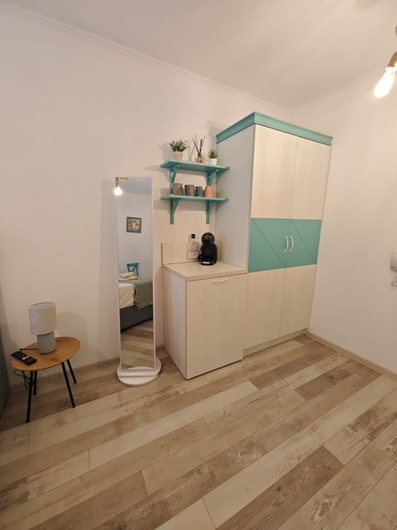 A small, minimalist room with light wood flooring, a white cabinet with turquoise accents, a tall mirror, a small wooden side table with a lamp, and two green wall shelves holding plants and decor.