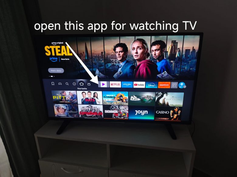 A TV screen displays a streaming app menu with various shows and apps. An arrow points to an app icon, and text above reads, open this app for watching TV. The TV sits on a white stand in a dimly lit room.