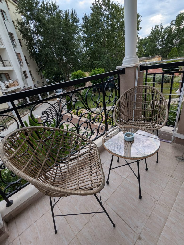 Two wicker chairs and a small round table with an ashtray on a balcony with a black railing. Trees and buildings are visible in the background.