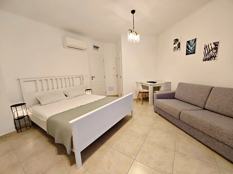 A bright, minimalist bedroom with a white bed, gray sofa, small dining table with two chairs, tiled floor, wall art, and overhead light fixture. The walls are white and the room feels spacious and tidy.