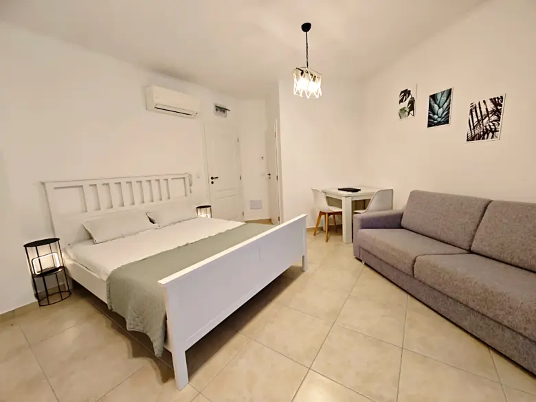 A bright, minimalist bedroom with a white bed, gray sofa, small dining table with two chairs, tiled floor, wall art, and overhead light fixture. The walls are white and the room feels spacious and tidy.