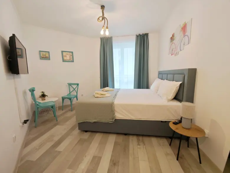 A modern bedroom with a double bed, light gray bedding, teal chairs, a small round table, framed wall art, light wood flooring, and large window with teal curtains. A TV is mounted on the wall.