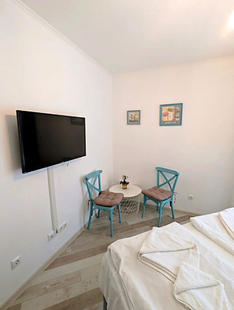 A bright, minimal room with a wall-mounted flat-screen TV, two blue chairs and a small round table, two framed pictures on the wall, and a neatly made bed with white linens in the foreground.