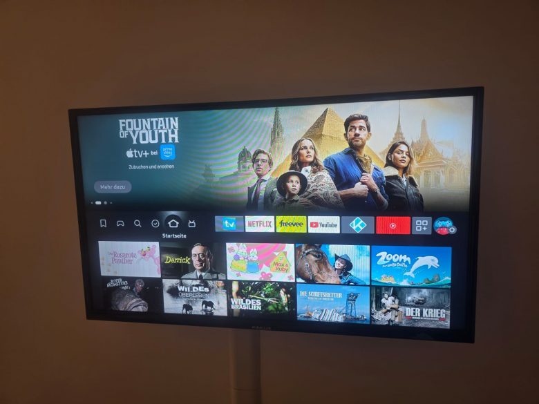 Televízor zobrazuje ponuku streamovacej služby, v ktorej je zvýraznený seriál Fontána mladosti s piatimi postavami na obrazovke. Pod hlavným výberom sa v riadkoch zobrazujú rôzne ikony aplikácií a miniatúry seriálov.