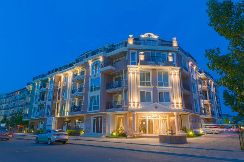 A modern, multi-story apartment building with illuminated exterior lights at dusk, featuring balconies and large windows; cars are parked along the street and trees line the sidewalk.