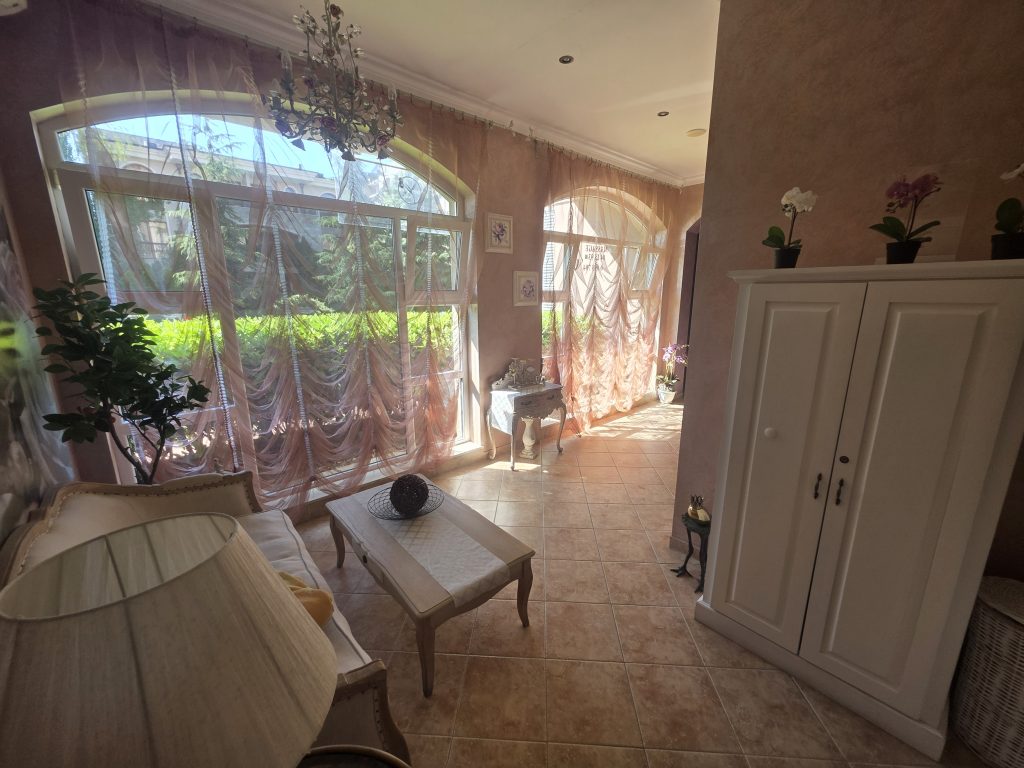 Bright sunlit room with large arched windows covered by sheer curtains, white furniture including a wardrobe and a table, tiled floor, houseplants, and decorative items creating a cozy, elegant atmosphere.