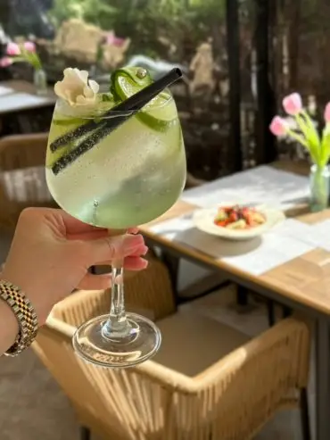 A hand holds a large wine glass filled with a green cocktail, garnished with a white flower, cucumber slices, and a black straw. Sunlight streams over a table with flowers and food in the background.
