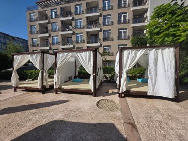 Three outdoor cabanas with white curtains and blue pillows are set in front of a multi-story apartment building, surrounded by greenery under a clear sky.