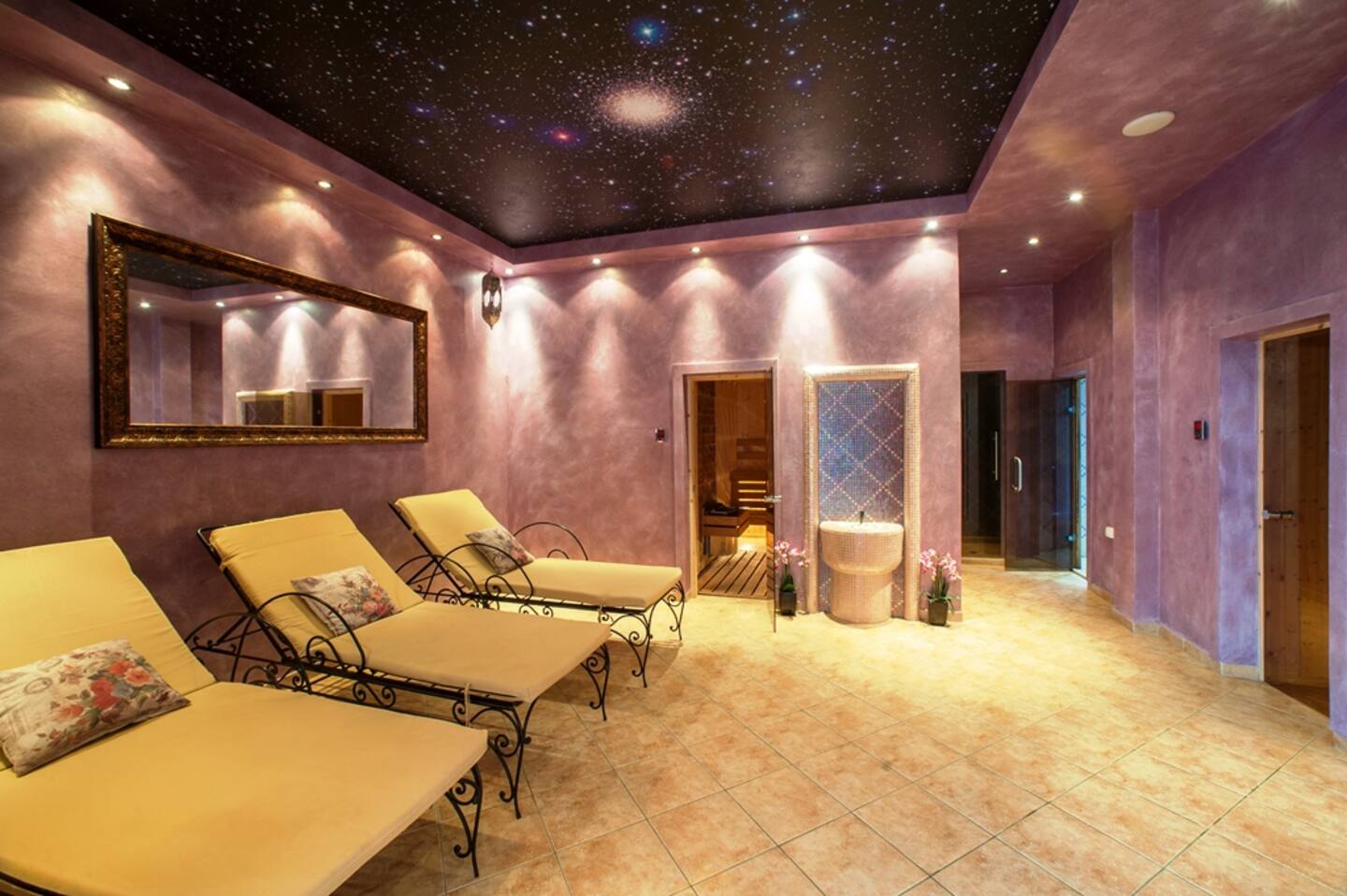 A spa relaxation room with three yellow lounge chairs, a large mirror, purple walls, a black starry ceiling, soft lighting, and open doors leading to a sauna and tiled shower area.