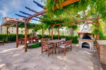 Outdoor patio with wooden pergola covered in green vines, wooden tables and chairs, and a stone barbecue grill. The area is surrounded by greenery and trees under a blue sky.
