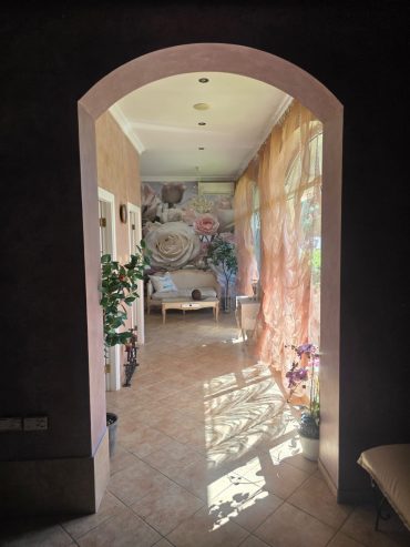 Sunlight streams through sheer curtains into a hallway with tiled floors, potted plants, and a mural of a reclining woman on the far wall. The space feels warm and inviting.