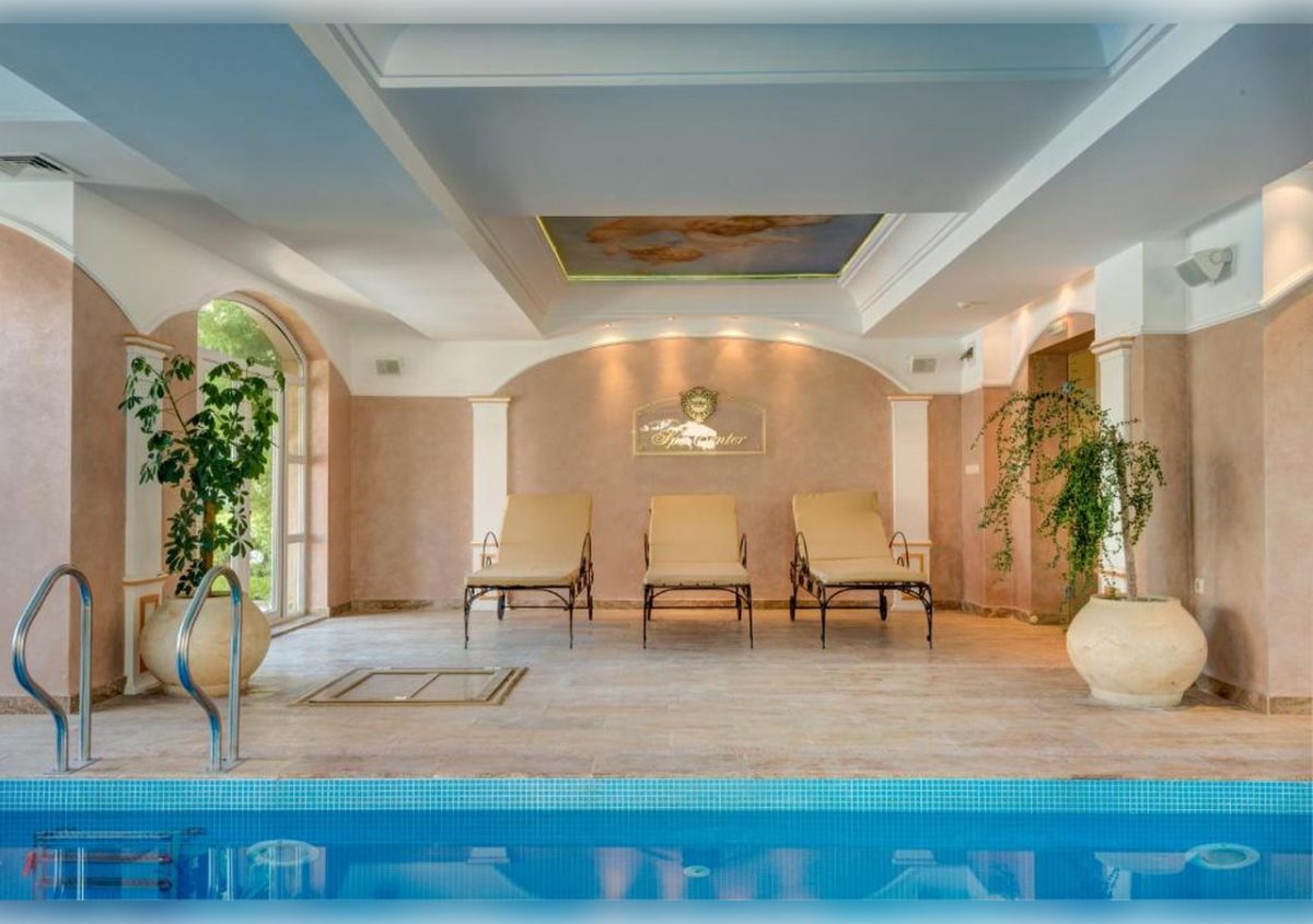 An indoor pool area with three beige lounge chairs against a tan wall, large potted plants on either side, arched windows, and a ceiling mural. The scene is calm and elegantly decorated.