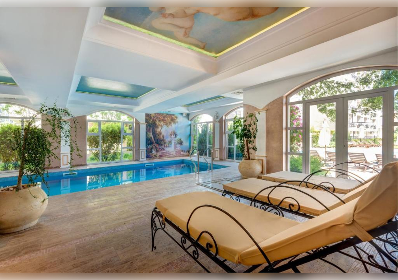 Indoor pool area with large windows, greenery, and two cushioned lounge chairs. The ceiling is decorated with murals and the sun shines through tall windows, illuminating the relaxing space.
