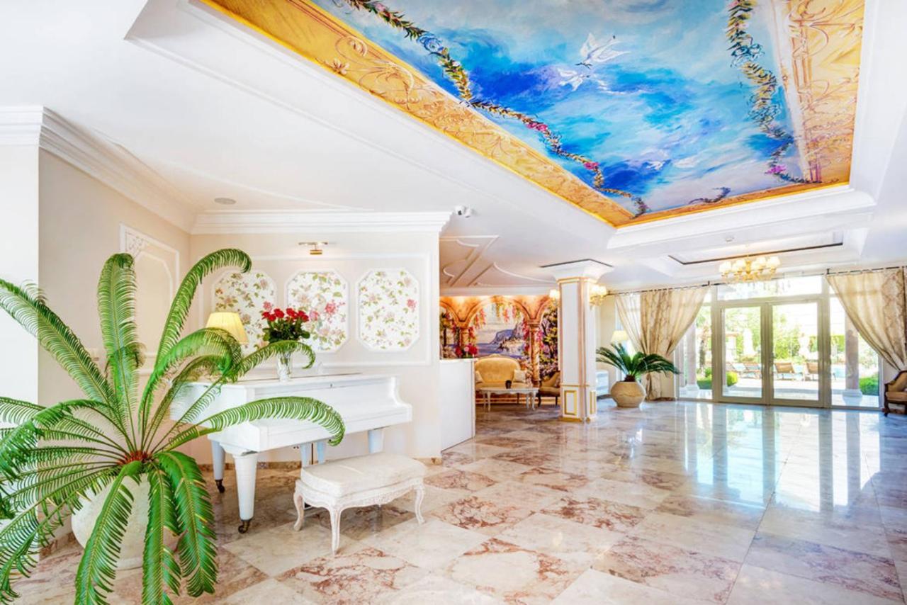 Eine luxuriöse, lichtdurchflutete Lobby mit Marmorböden, einem weißen Flügel, großen Topfpflanzen und einer Decke, die mit einer lebhaften Himmelslandschaft bemalt ist. Vom Boden bis zur Decke reichende Fenster lassen viel natürliches Licht herein.