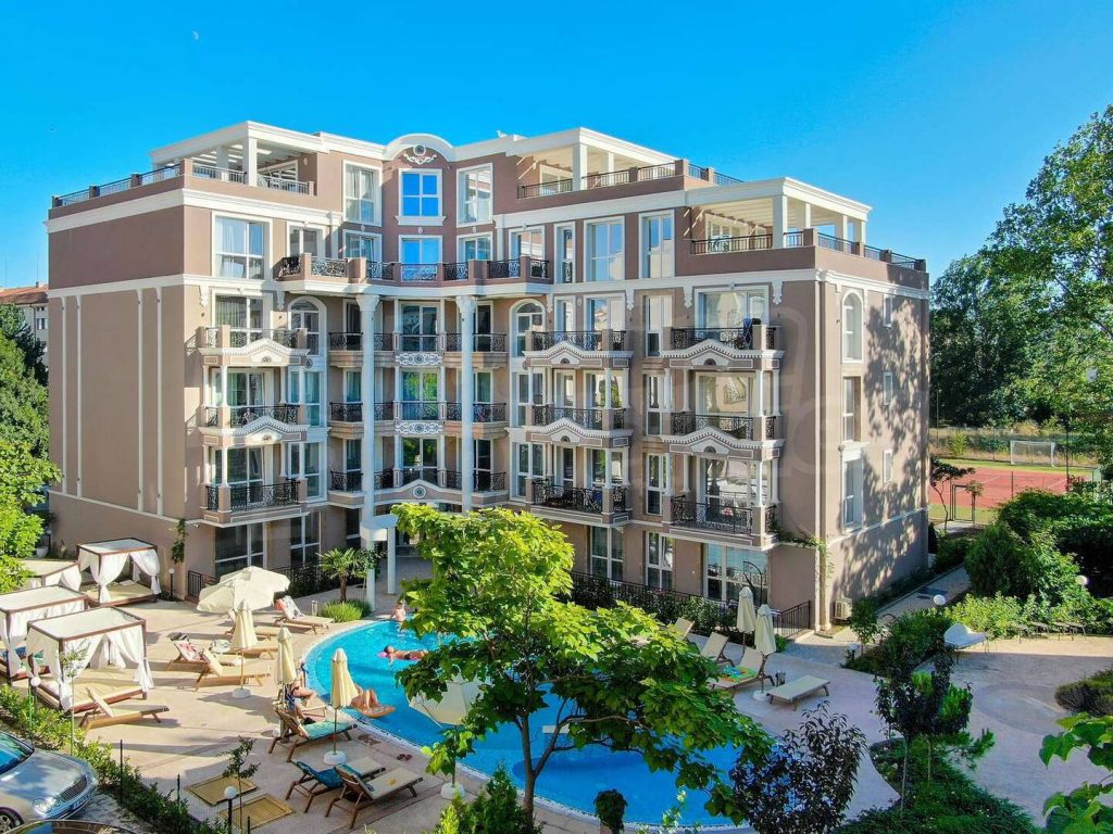 A multi-story hotel with balconies overlooks a courtyard featuring a swimming pool, sun loungers, and umbrellas. Lush greenery surrounds the area, and the sky is clear and blue.