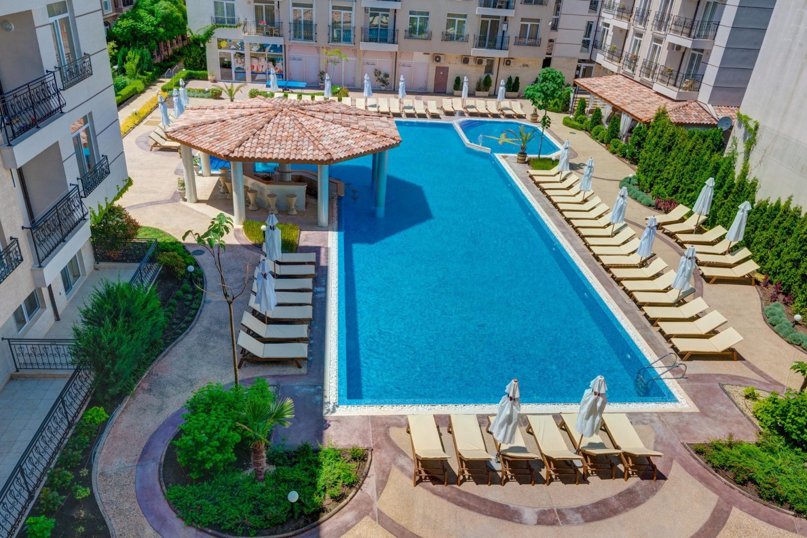 A rectangular outdoor swimming pool surrounded by lounge chairs with umbrellas, bordered by landscaped greenery and multi-story buildings in the background.