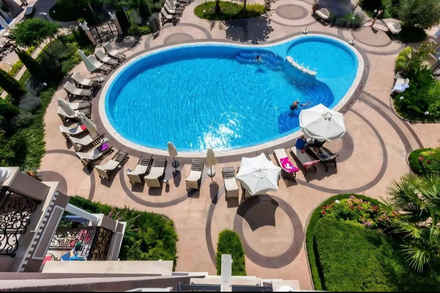 Aerial view of an outdoor swimming pool with a unique shape, surrounded by lounge chairs, sun umbrellas, and landscaped gardens. Several people are relaxing by the pool and enjoying the sunny weather.