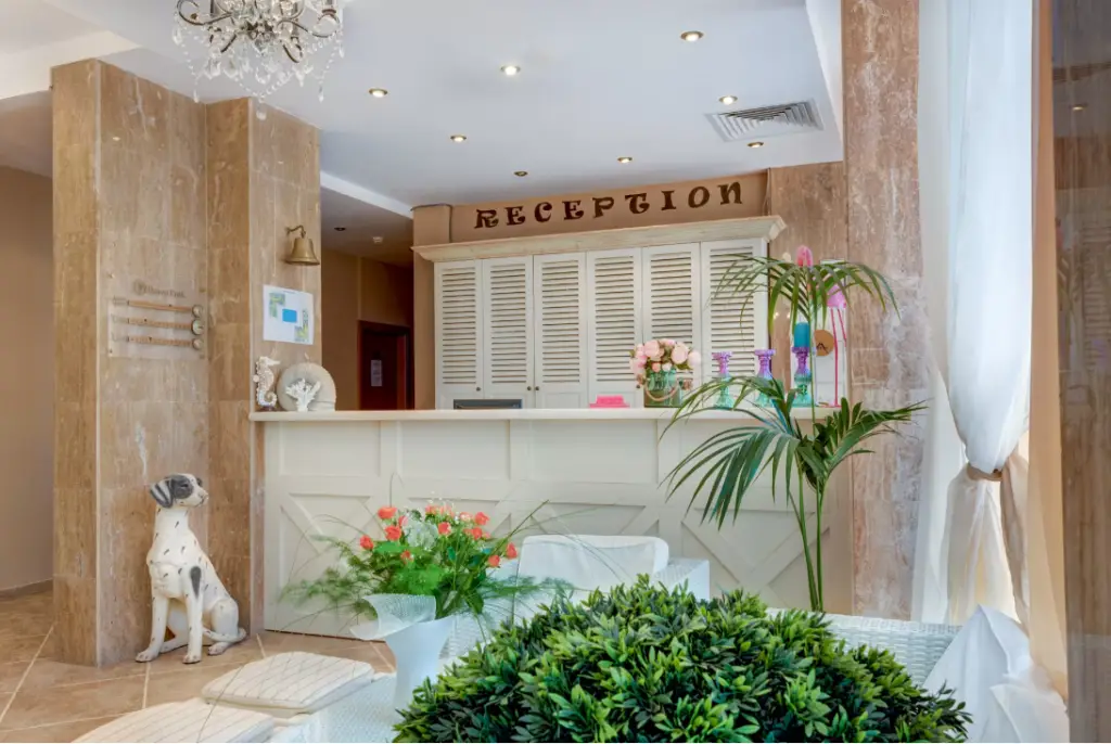 Hotel reception area with a white front desk, flowers, potted plants, a dog statue, and a chandelier. The word RECEPTION is displayed on a wooden sign above the desk.