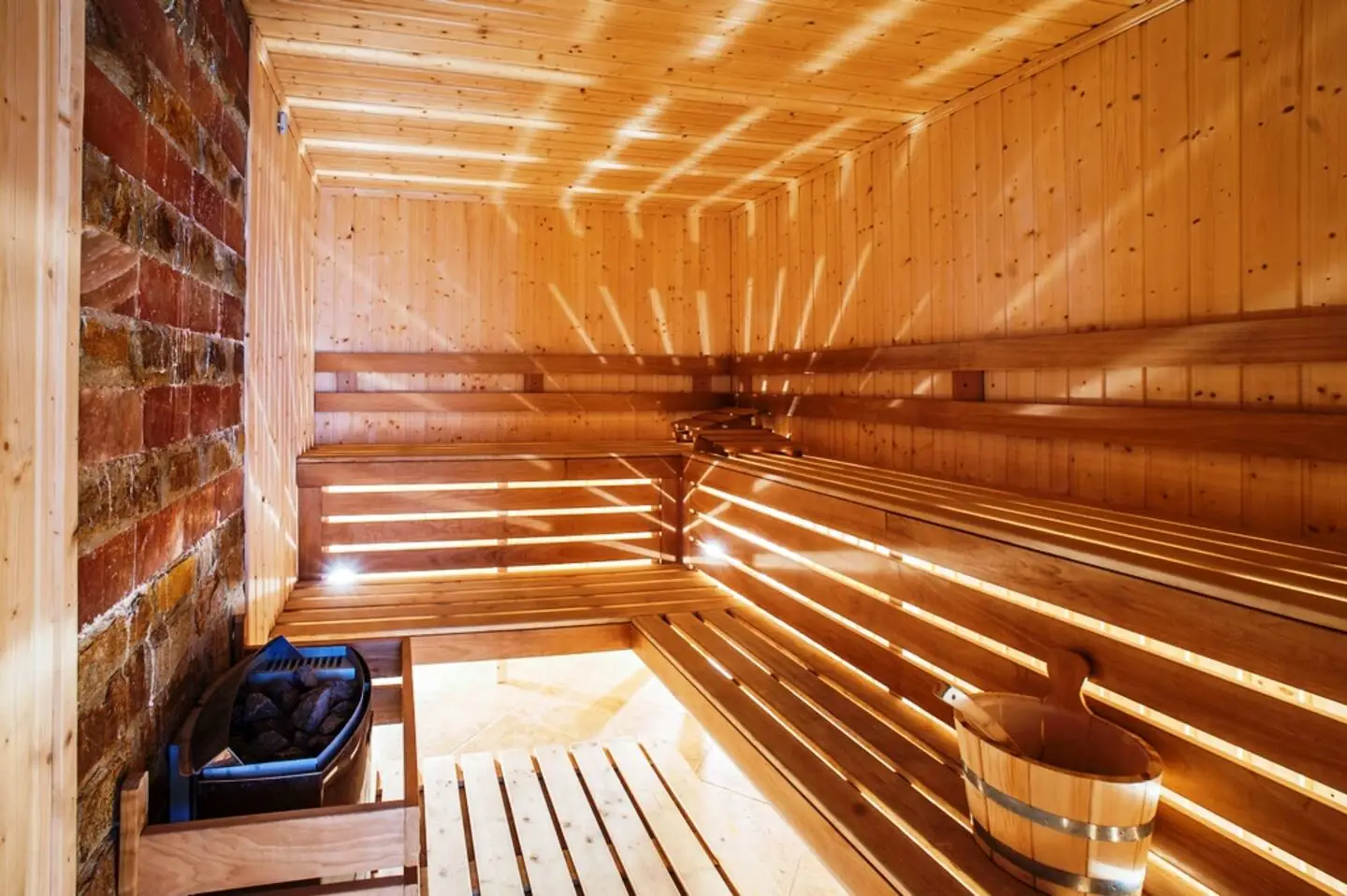 Eine geräumige Hotelsauna mit Holzbänken, Holzwänden und -decke, einem Eimer mit einer Schöpfkelle und einem Steinofen in einer Ecke. Warmes Licht erzeugt Muster auf den Holzoberflächen.