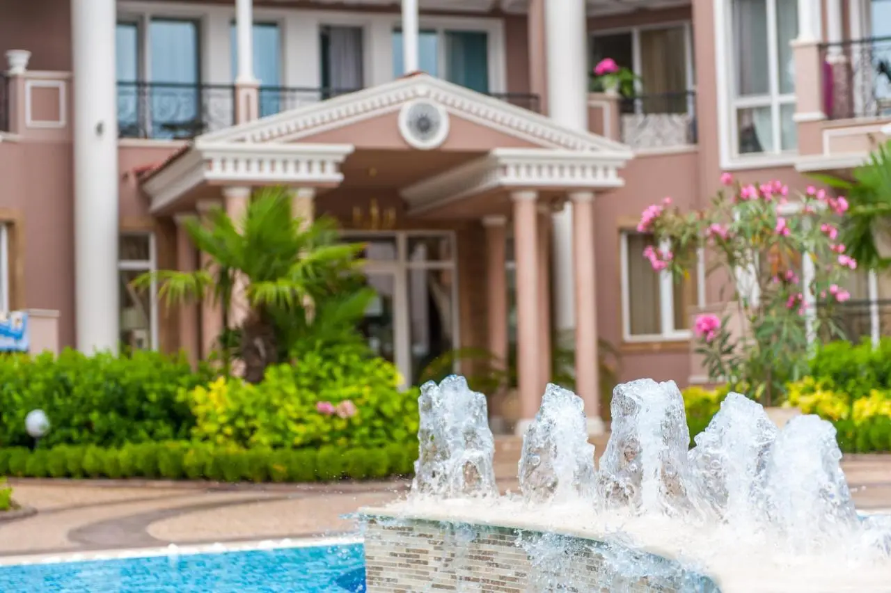 A close-up of a water fountain with streams of water rising, set in front of a large hotel or mansion with columns, balconies, lush greenery, and blooming flowers.
