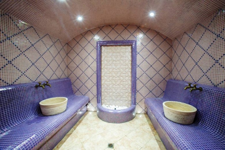 A tiled hotel steam room with curved walls and ceiling, purple and pink mosaic benches, two stone washbasins, and a decorative fountain at the centre of the back wall. Bright lights illuminate the inviting space.