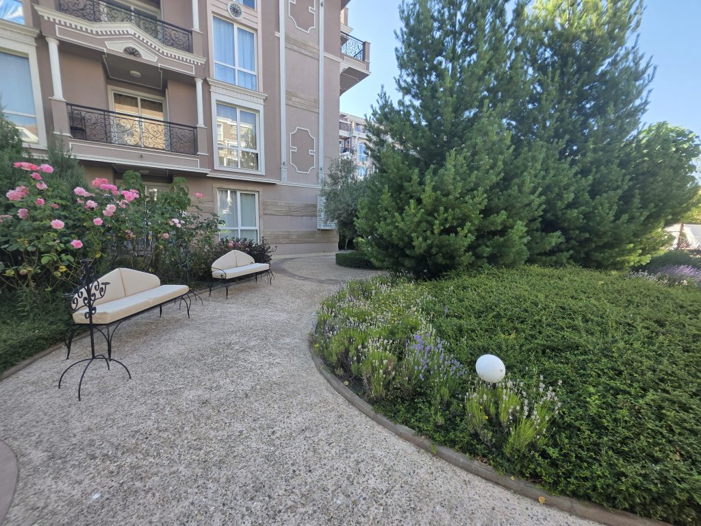 A curved path with two white benches winds through a garden of flowering plants and green shrubs, beside a beige hotel building with balconies. A round white garden lamp stands in the foreground.