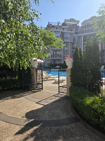 A sunlit hotel courtyard with an open wrought-iron gate leads to a pool surrounded by loungers, lush greenery, and elegant multi-storey buildings in the background.