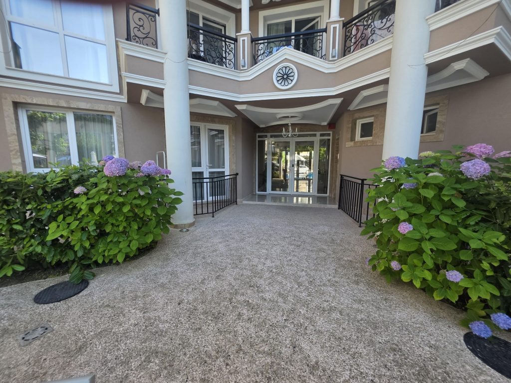Entrance to a hotel building with two large white columns, glass doors, beige walls, black railings on the balcony above, and blooming hydrangea bushes lining the path.