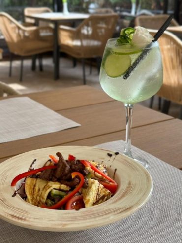 A plate of food with grilled meat, red peppers, and vegetables sits on a table next to a tall glass of a clear drink garnished with cucumber, lime, and a white flower, highlighting the dining amenities available amid wicker chairs in the background.