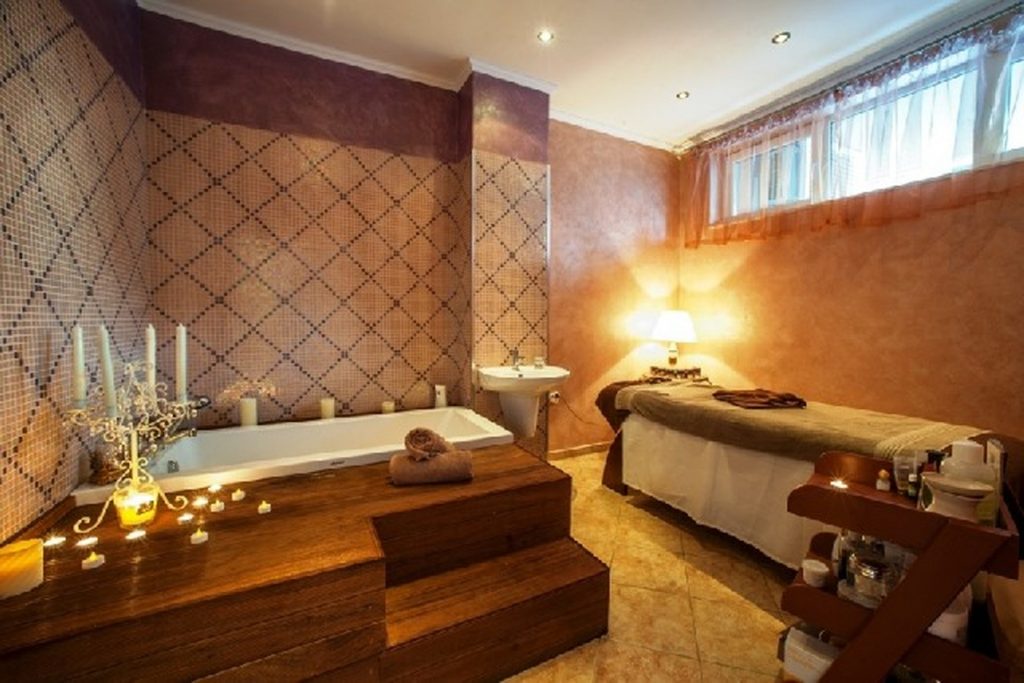 A cozy spa room with a bathtub surrounded by candles, a massage table with towels, warm lighting, and tiled walls. A small sink and a window with curtains are in the background.