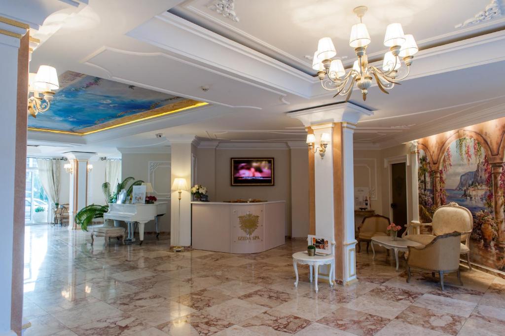 Elegant hotel lobby with marble floors, a chandelier, a white grand piano, plush armchairs, a reception desk with a TV above it, potted plants, and decorative wall art and ceiling mural. Bright and inviting atmosphere.