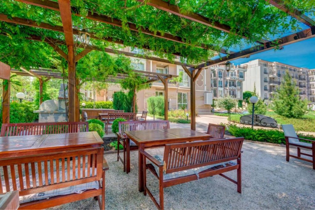 Wooden tables and benches sit under a leafy pergola in a hotel's outdoor garden area, surrounded by greenery and blocks of flats beneath a clear blue sky.