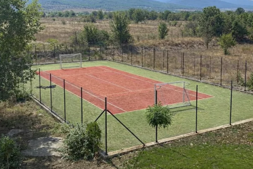A small outdoor sports court with amenities like a red tennis surface, football goals on green turf, all enclosed by a tall wire fence, set in a rural area with dry grass and scattered trees.