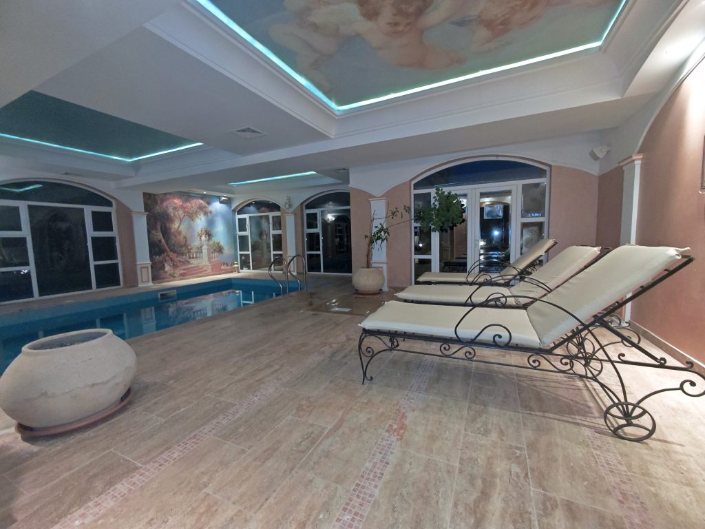 Indoor pool area with two cushioned lounge chairs, large windows, and a painted ceiling. The floor is tiled, and there is a decorative vase on the left. The setting appears calm and luxurious.