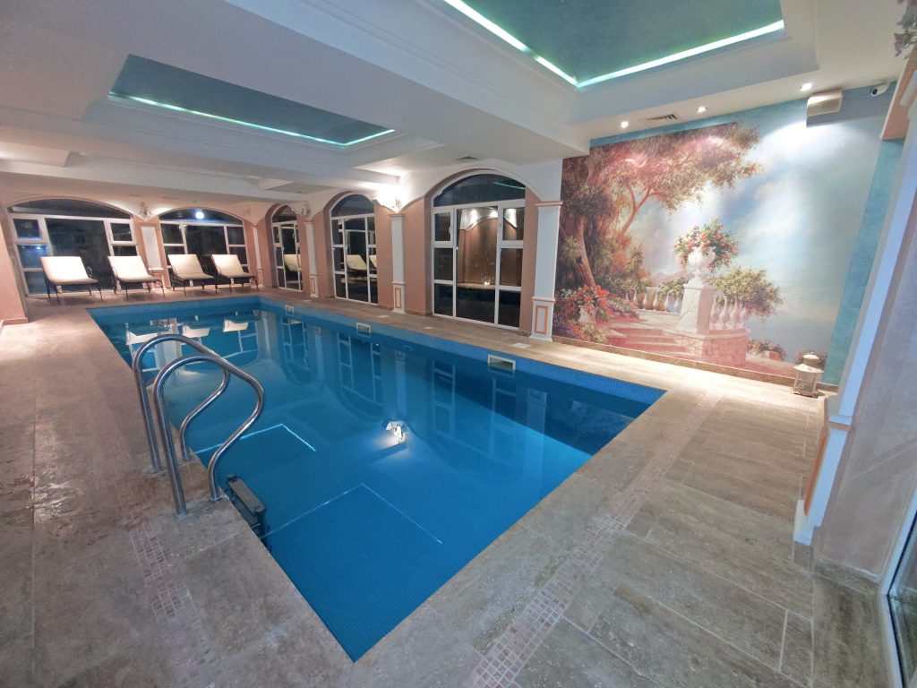 Indoor swimming pool with clear blue water, surrounded by lounge chairs and large windows. The wall features a mural of a scenic outdoor garden with steps, trees, and flowers. The area is well-lit with ceiling lights.