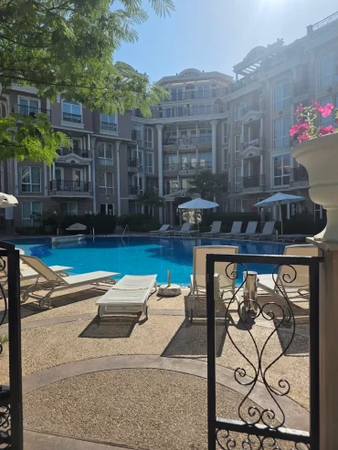 A view through an open gate reveals a sunny courtyard with a swimming pool, surrounded by lounge chairs and umbrellas, and a multi-story apartment building in the background. A potted plant with pink flowers sits on the right side.