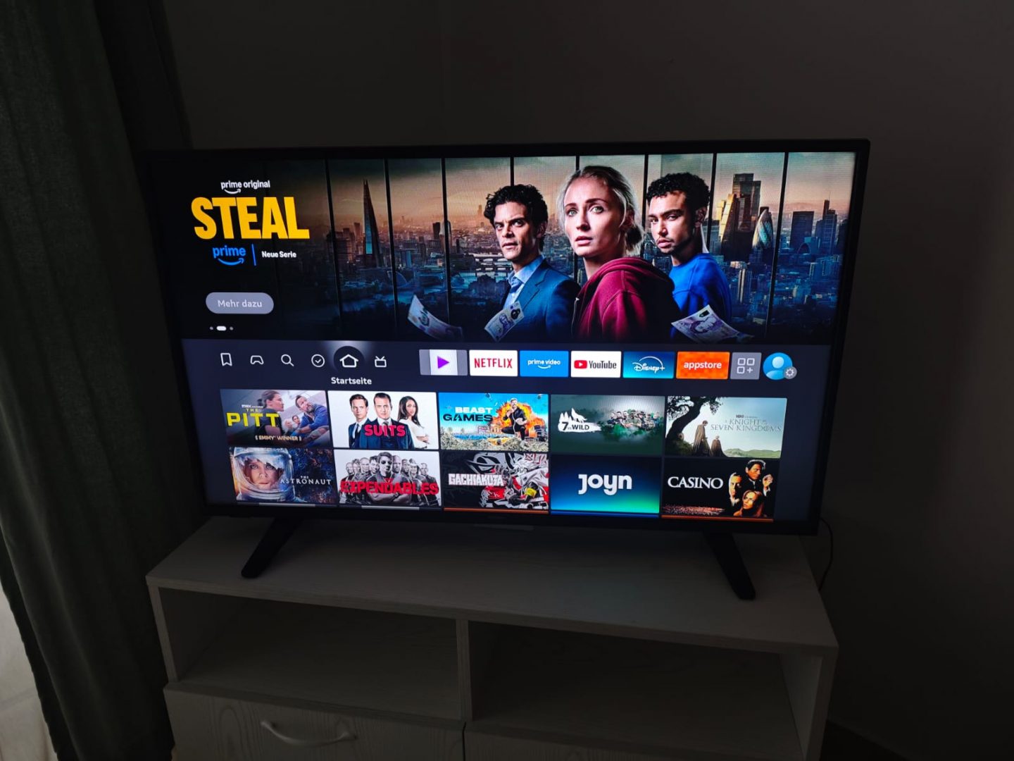 A flat-screen TV on a white stand displays a streaming service menu, highlighting the show Steal with three people holding money. Various app icons like Netflix, Prime Video, and Disney+ appear below.
